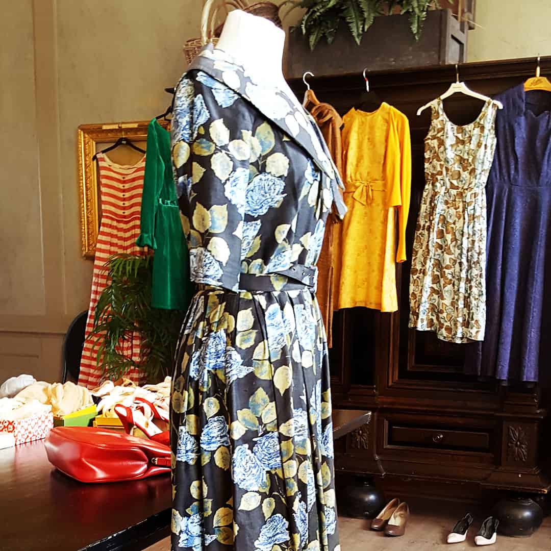 Mannekäng i blå blommig klänning i en vintagebutik, omgiven av klänningar och skor i en stilfull miljö.