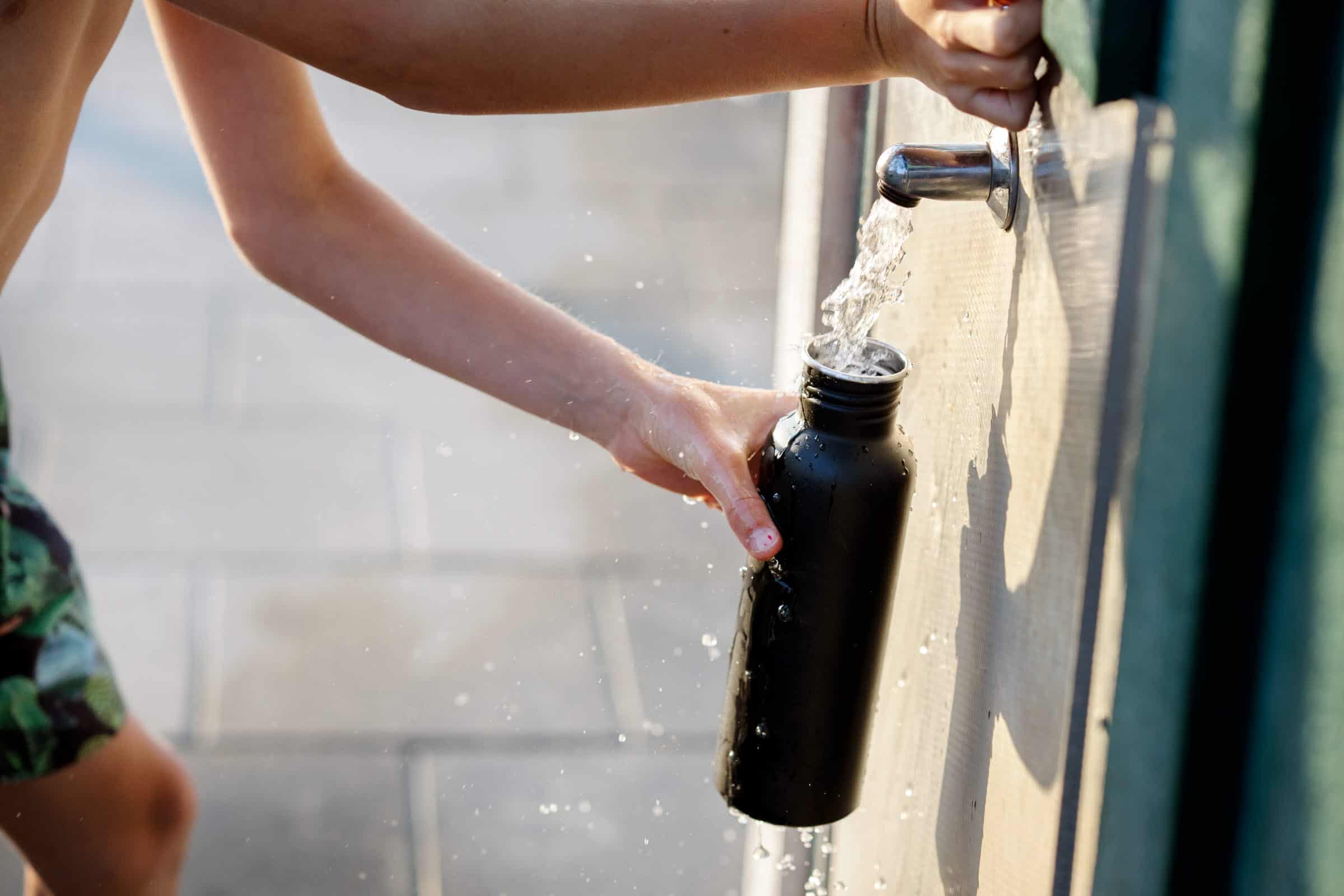 Vid en vattenpost på en vägg fylls en svart vattenflaska. Vattnet porlar och stänker ner handen som håller i den.