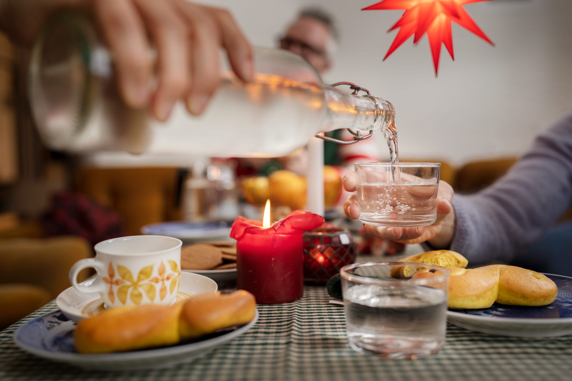 I ett ljust rum över ett bord med smårutig grön duk lyser en flertaggig rödgul stjärna. På duken står ett glas vatten och tända ljus i olika röda nyanser. Bordet är dukat med kaffekoppar och assietter. På assietterna ligger lussekatter och pepparkakor. Vid bordets bortre kortända skymtar apelsiner och en man. Någon sträcker sig över bordet med en gammeldags klar flaska med vatten. Från andra sidan bordet håller ett barn fram ett glas. Vattnet bubblar när det hälls i glaset från flaskan.